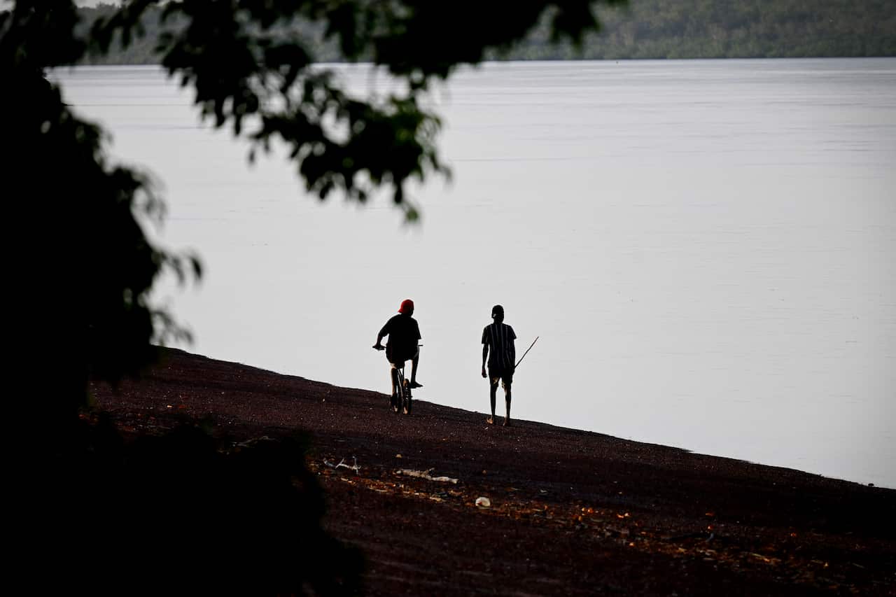 Silhouette of two people on the shore next to water. One is holding a long spear and the other riding a bike.