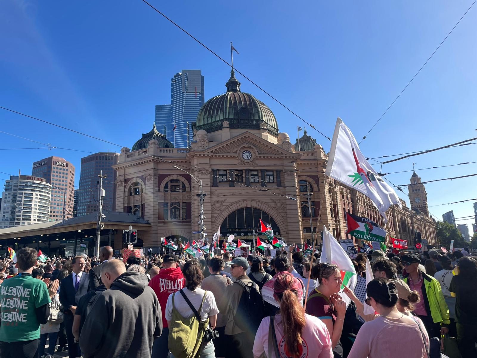 A large crowd of people carrying Palestinian flags and banners gathers for a protest in front of Flinders Street Station.