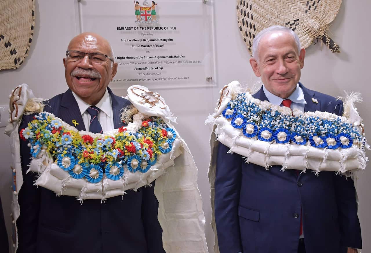 Two men in dark suits wearing large, elaborate ceremonial flower garlands stand side-by-side smiling in front of an official wall plaque.