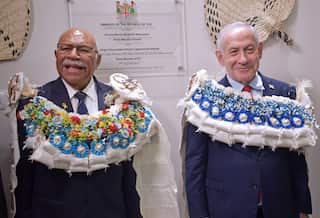 Two men in dark suits wearing large, elaborate ceremonial flower garlands stand side-by-side smiling in front of an official wall plaque.