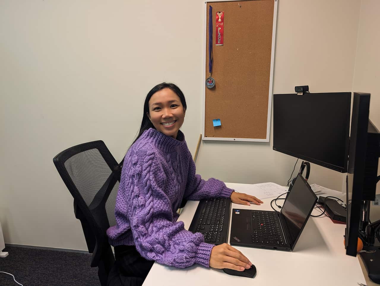An Asian woman wearing a purple jumper sits at a desk in front of a computer