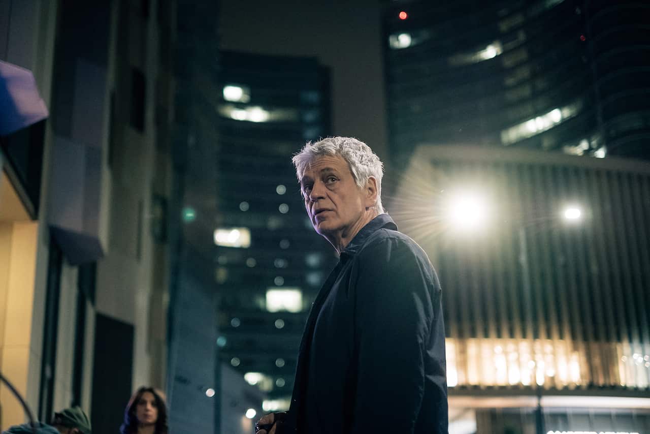 A grey-haired man in a dark coat stands in a city street at night, looking sideways.