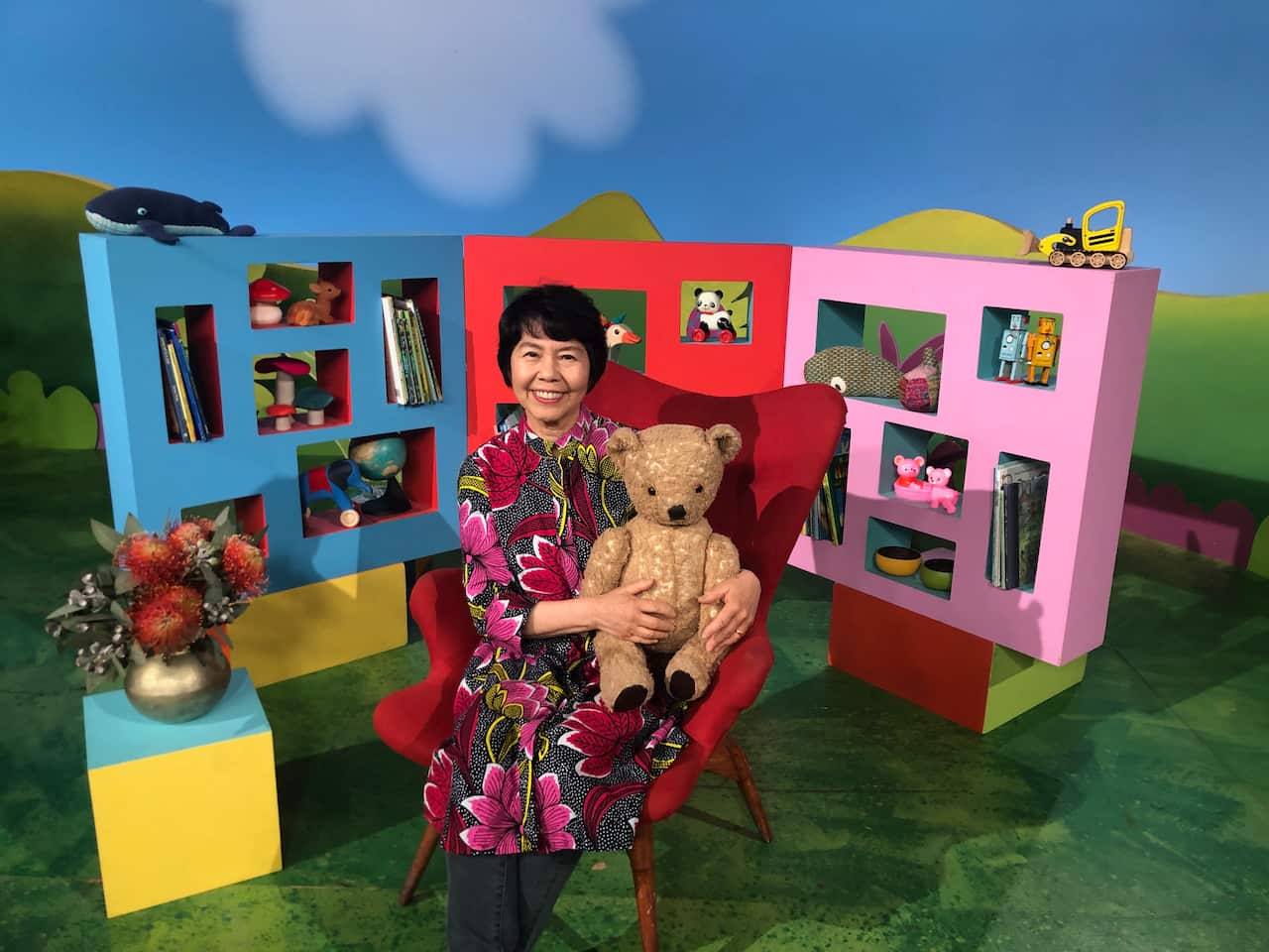 A woman on the set of Play School sitting in a red chair and holding a teddy bear. There are coloured bookshelves behind her.