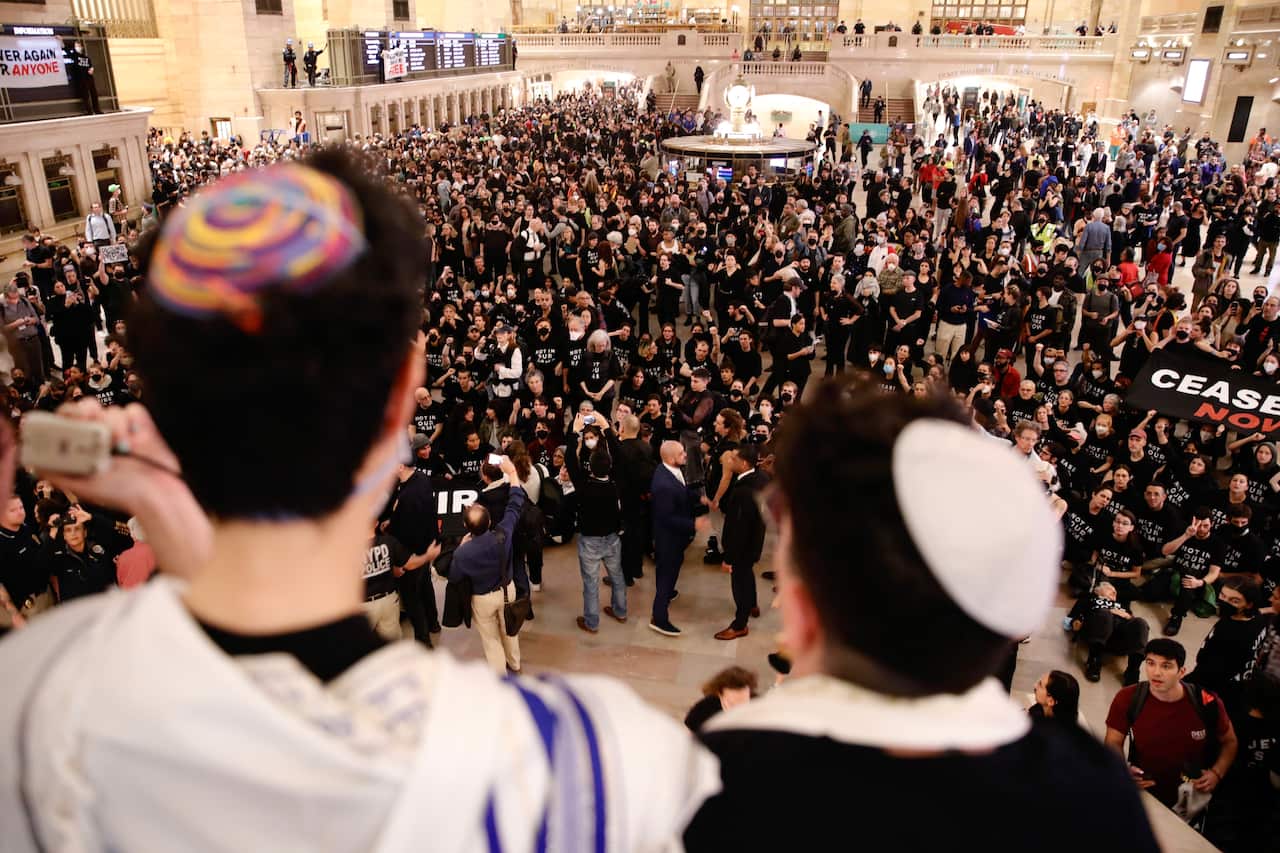 Two men wearing Jewish caps (kippot) in the foreground, looking down at crowds of people gathered for a protest in Grand Central Station.