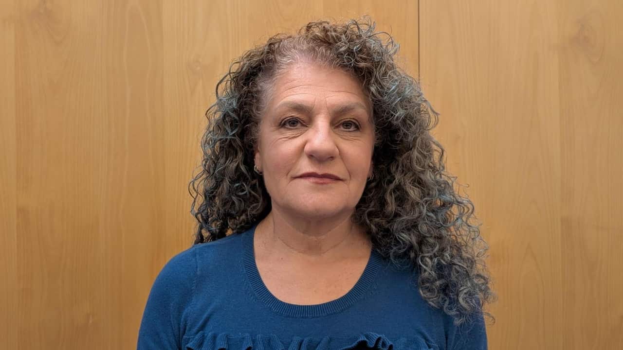 a closeup of a middle aged woman with grey curly hair in a blue top in front of a wooden background