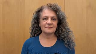a closeup of a middle aged woman with grey curly hair in a blue top in front of a wooden background