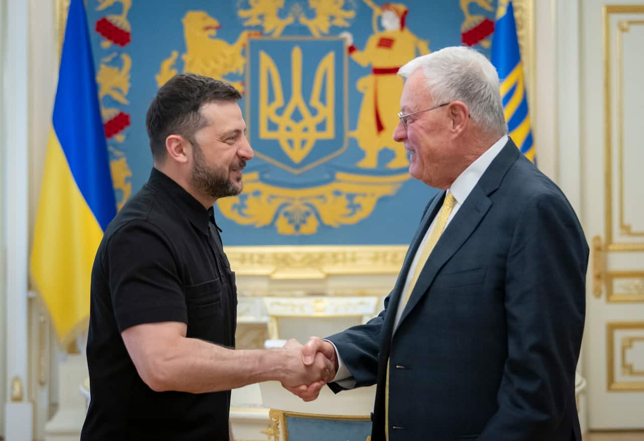 Two men shake hands in front of Ukrainian flags, one wearing all black and the other in a suit.