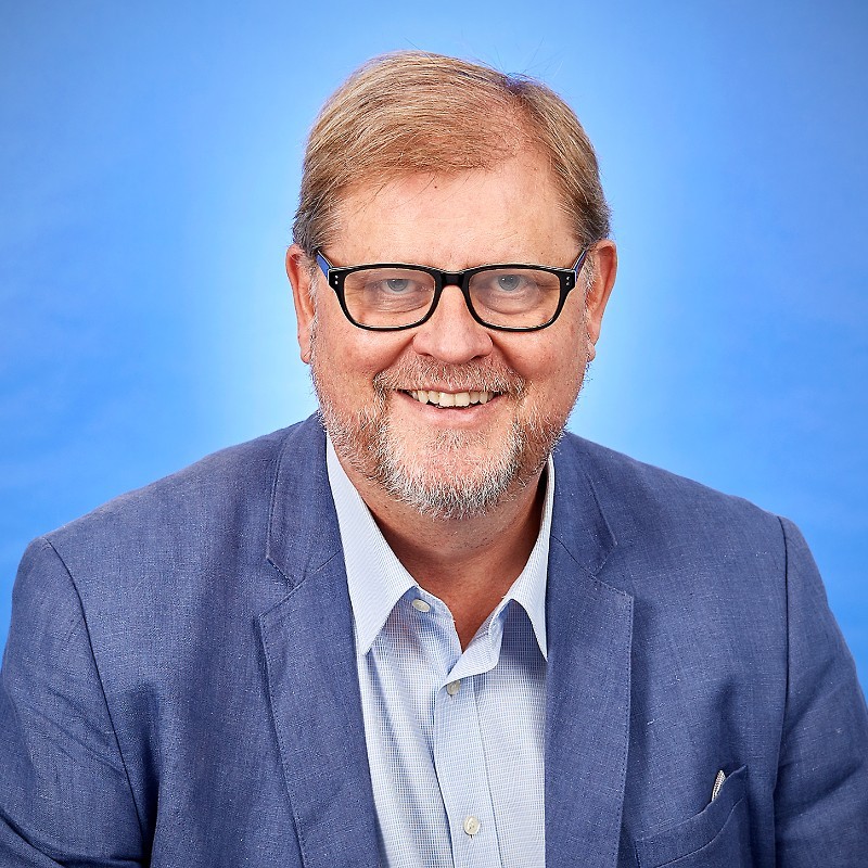 A man in a blue suit and black glasses in front of a blue background