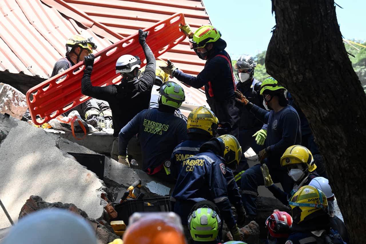 Rescue workers wearing dark uniforms and light-coloured helmets stand on top of a pile of rubble next to the trunk of a tree. The two at the top are carrying a red stretcher.