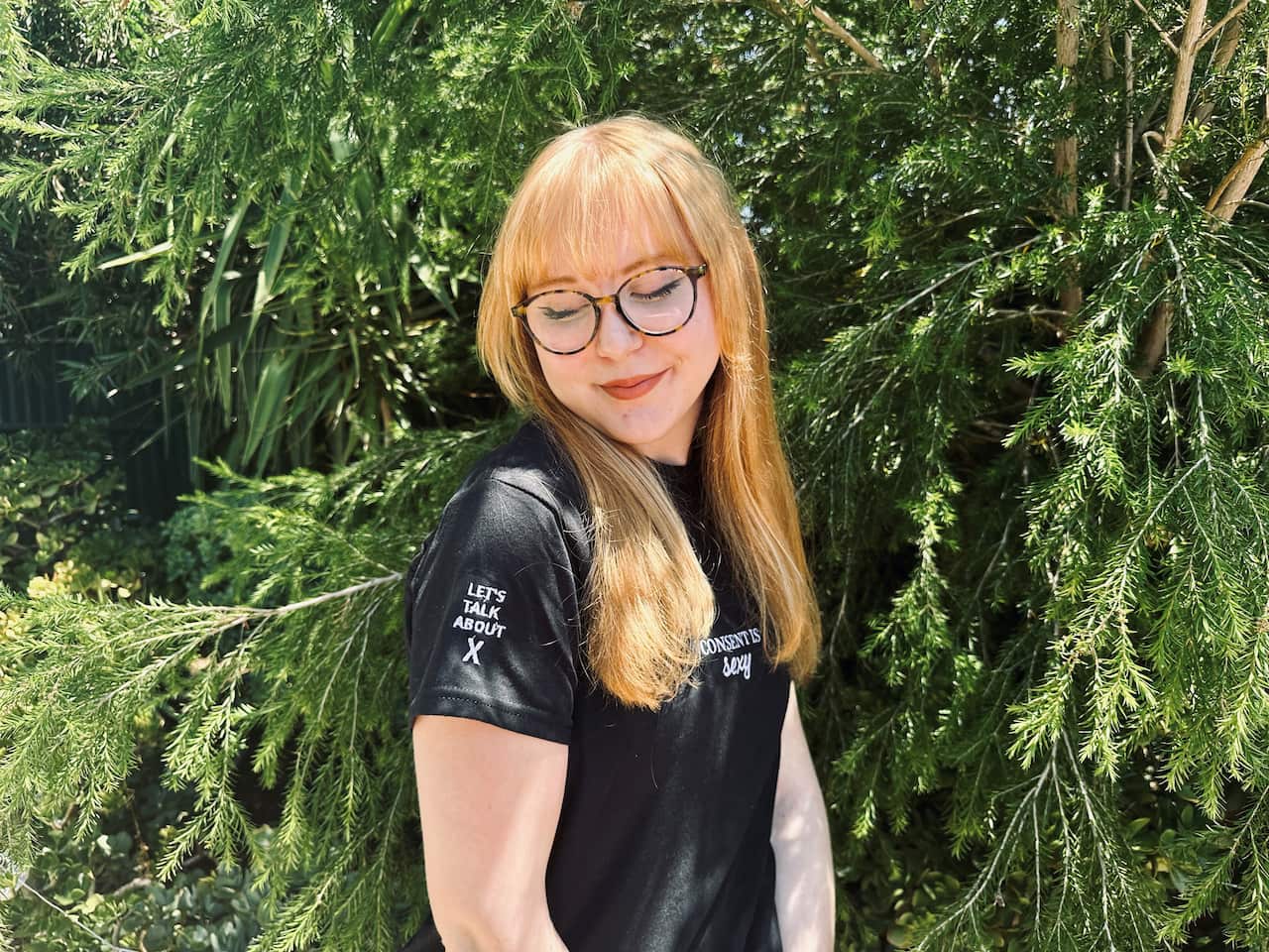 A young girl wearing glasses and a black t-shirt is smiling and looking down.