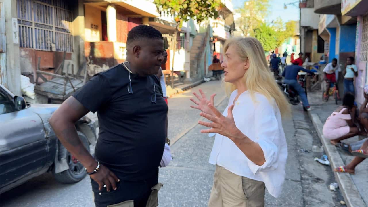 A man in a black T-shirt and pants stands opposite a woman with blonde hair, who's wearing a white shirt and khaki pants. She is gesticulating as she speaks. Rundown buildings, a burnt car, and people are visible in the background.  