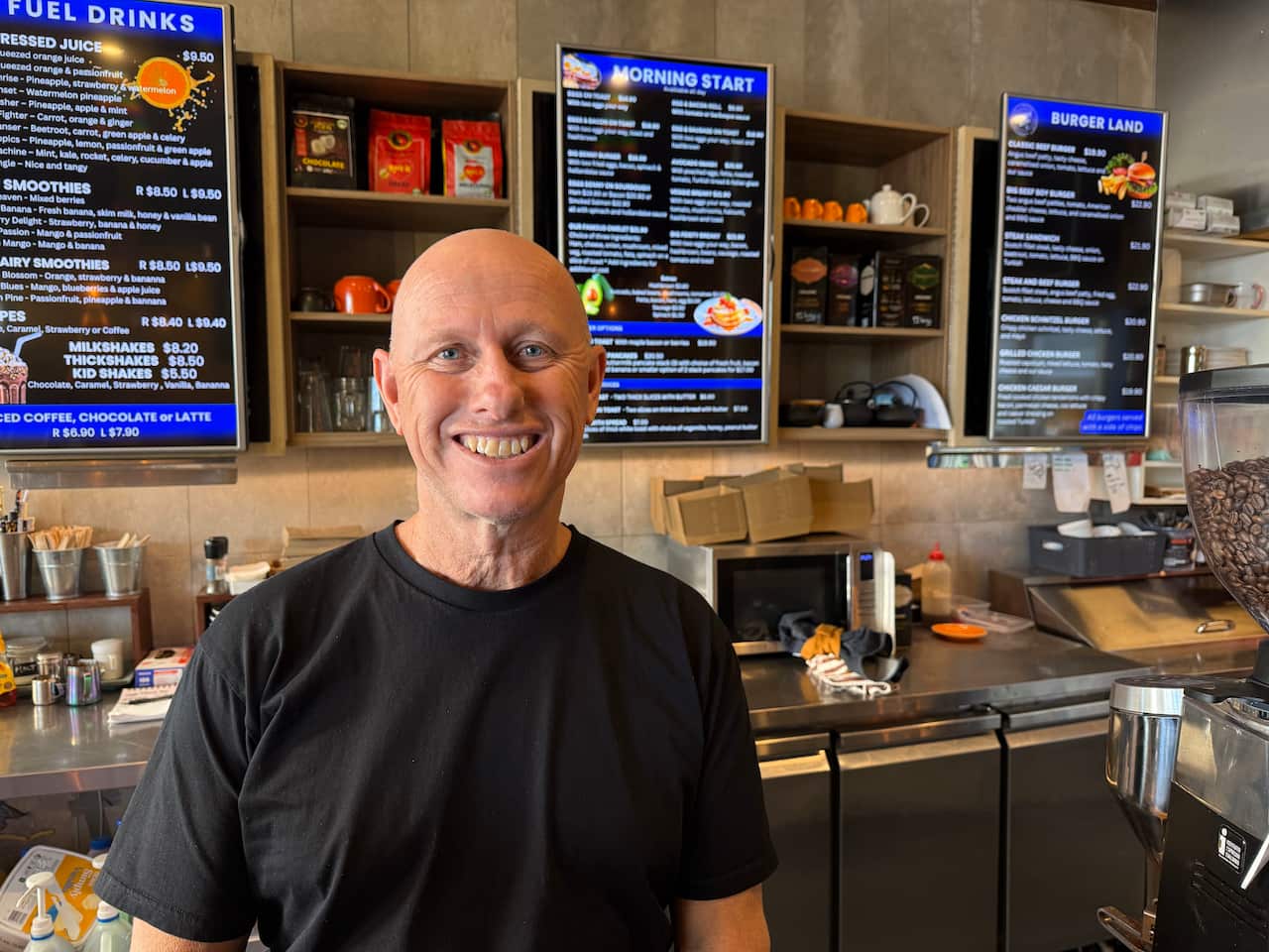 A man wearing a black t-shirt stands in front of a cafe menu