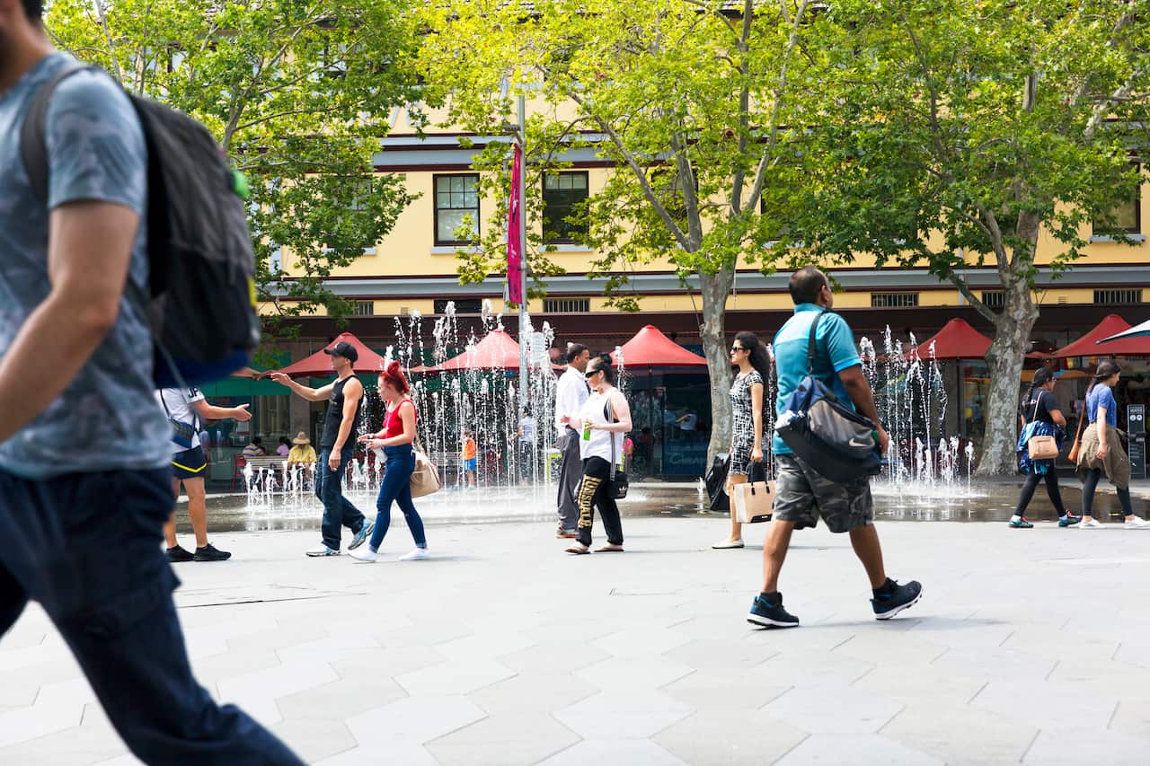 People walking through a city plaza with fountains and red umbrellas