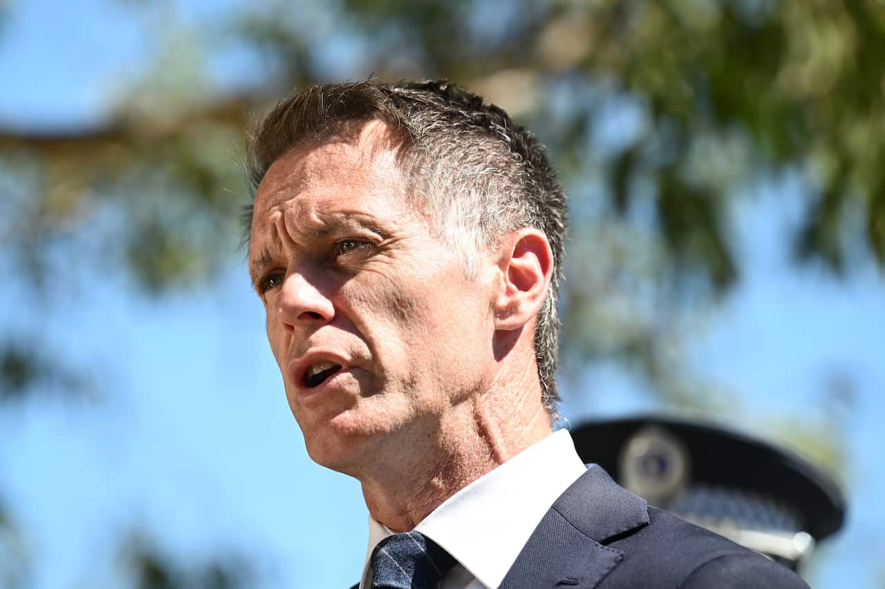 Chris Minns, wearing a dark suit and white shirt, speaking while outside. A police officer's hat is visible behind him.