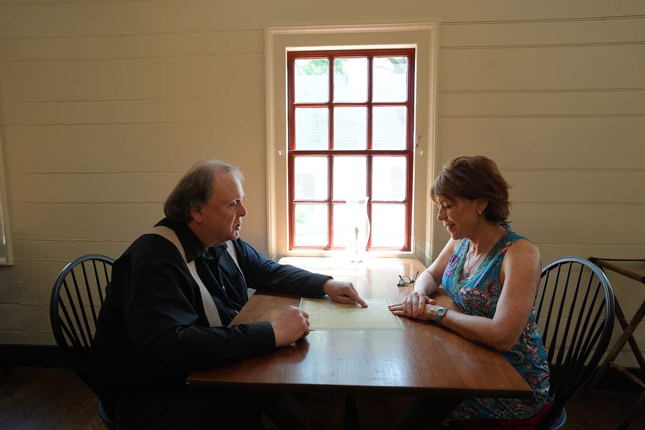 A smiling woman in a sleevless patterned dress and a man in a dark shirt and braces sit on either side of a wooden table. He is pointing to piece of paper on the table, as if explaining something. 