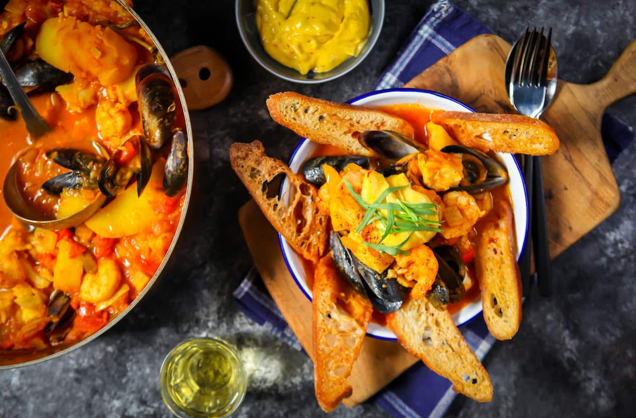 Seen from overhead, a big pot of fish stew sits on one side. Beside it sits a plate with a serve of stew, toasted slices of bread and a dollop of yellow mayonnaise. A bowl of mayonnaise sits behind the plate. 