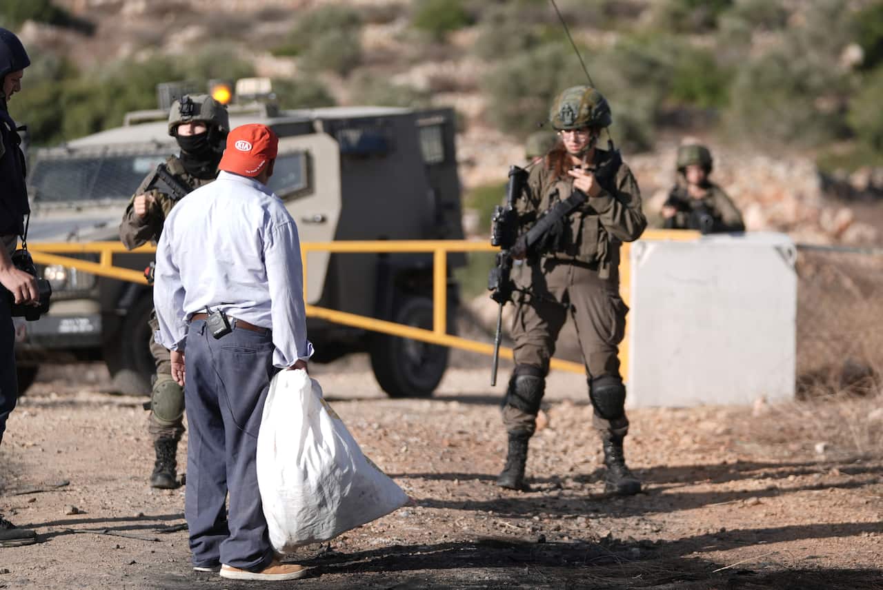 A man holding a large white bag, standing on a dirt road. He is surrounded by soldiers in green uniforms, carrying guns.