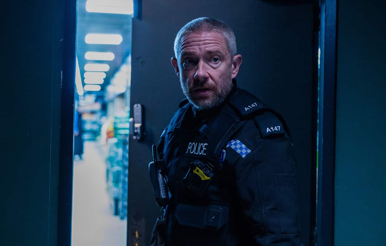 A man in a police uniform stands in a dark doorway, with a well-light rooom partly seen through behind him.