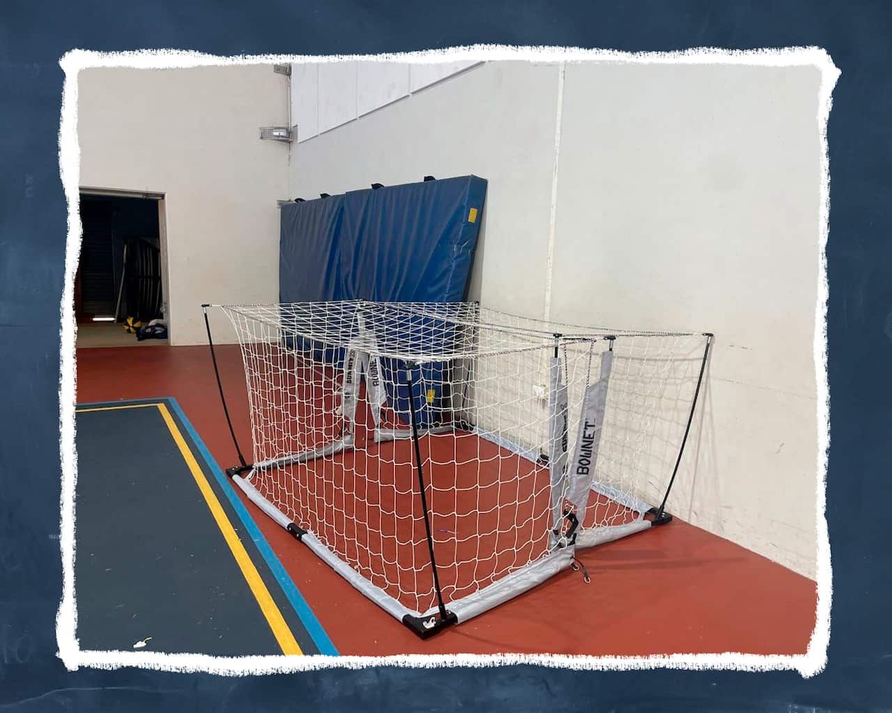 A makeshift cage constructed with two indoor hockey nets sits empty at the edge of an indoor sports facility.