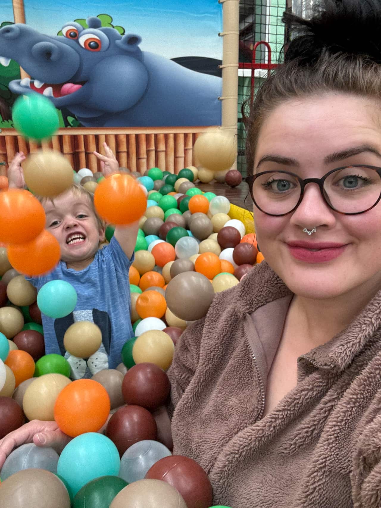 A selfie of a brunette woman with glasses and a young boy in a ball pit. The boy is throwing balls towards the camera and has an excited expression. 