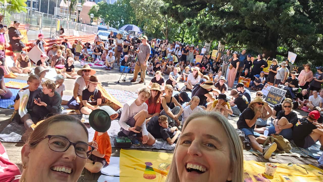 Two smiling women take a selfie in front of a large group of people sitting on picnic rugs.