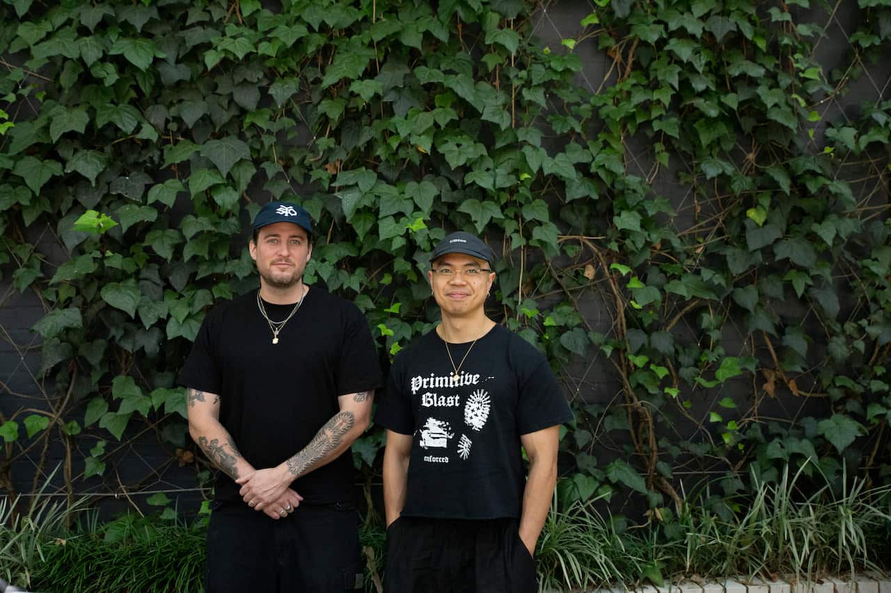 Two men wearing black smiling in front of a grassy wall.