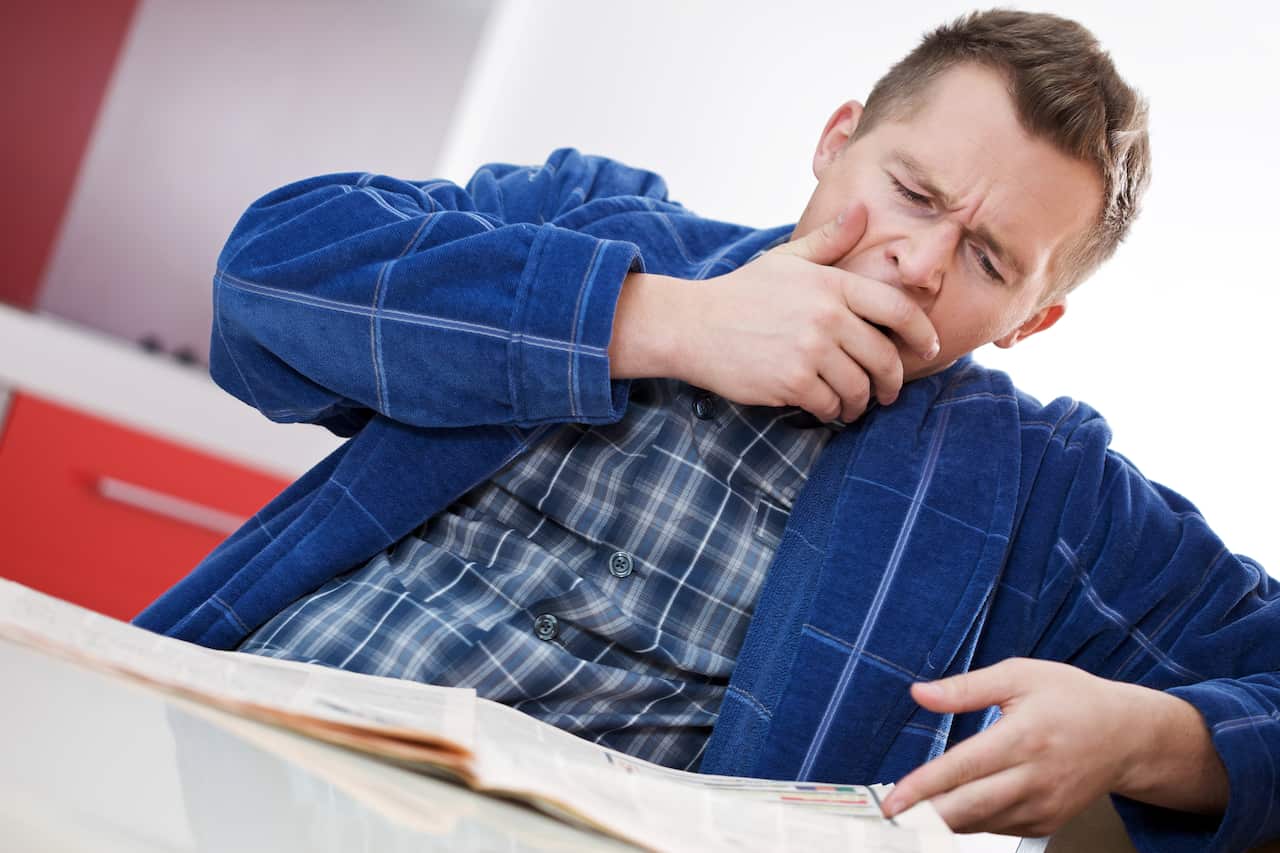 A young man sitting at a table with a book in front of him covers his mouth with his hand as he yawns