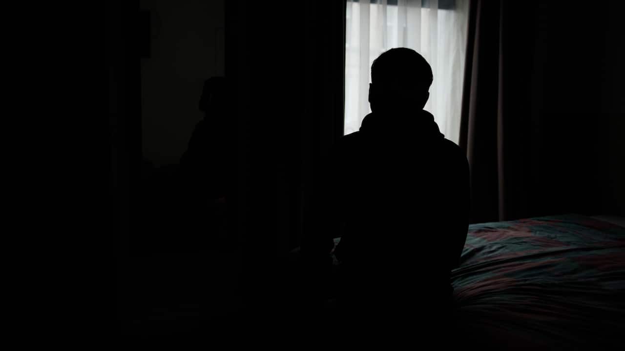 A shadowy image of an unidentified man sitting on a bed. Behind him is a window with the curtains drawn. 