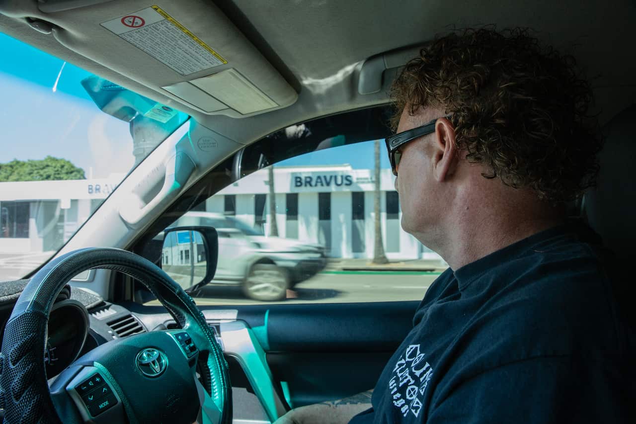 A middle-aged man wearing a black t-shirt drives a car past a large office building labelled 'Bravus'.