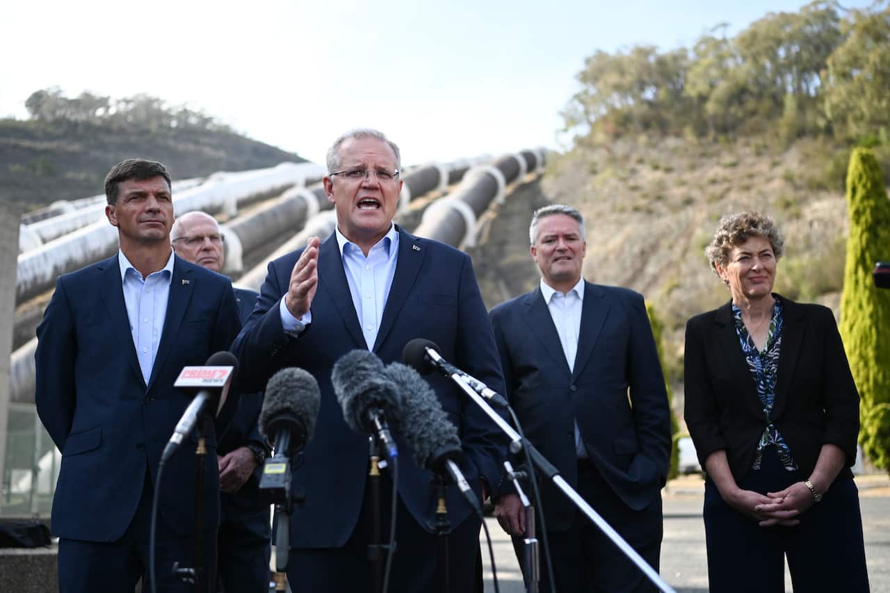 Former prime minister Scott Morrison, wearing a suit, speaks into microphones outside. There are three men and a woman in suit standing behind him, and in the background pipelines running up a hill