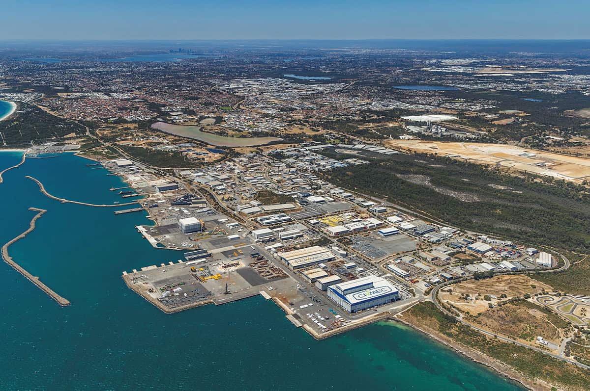 An aerial view of the Australian Marine Complex in Henderson, Western Australia, shows a large industrial port facility next to a body of water. In the background, a sprawling cityscape with multiple lakes is visible under a clear sky.
