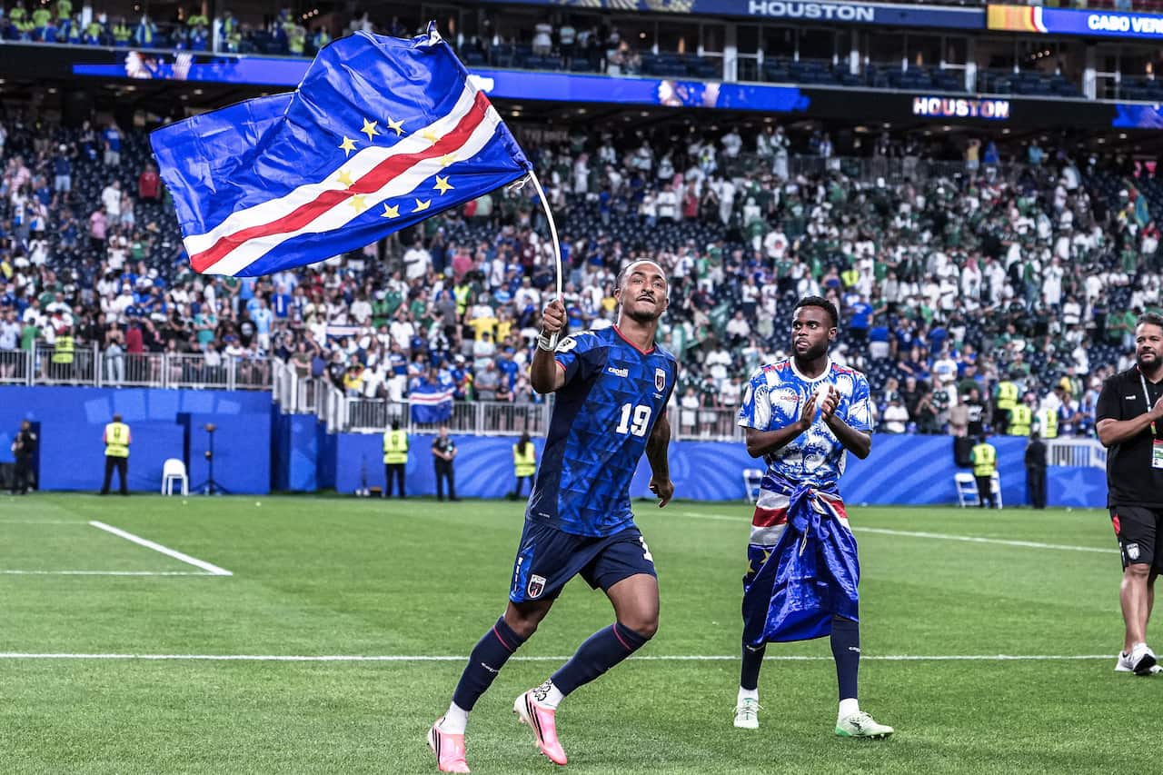 A footballer in blue shirt and shorts waves a blue, red and white flag on a stick while walking across the pitch after the match, as a teammate applauds nearby. Fans in the stands celebrate in the background.