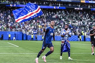 A footballer in blue shirt and shorts waves a blue, red and white flag on a stick while walking across the pitch after the match, as a teammate applauds nearby. Fans in the stands celebrate in the background.