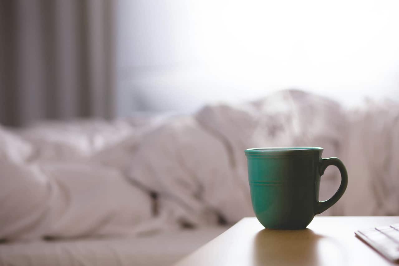 A dark green coffee mug is placed on the table with a bed in the background.