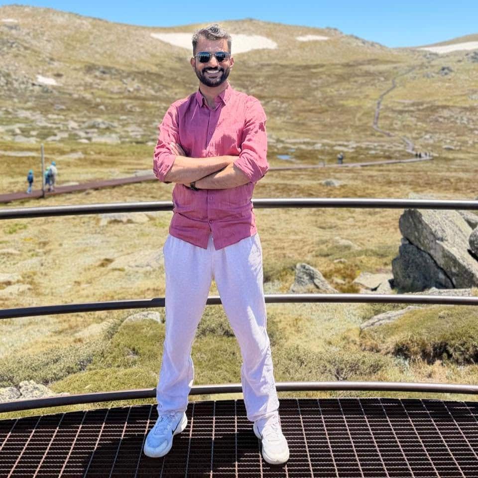 A man in a pink shirt and white pants stands with his arms folded on an iron platform with a dry, hilly terrain in the background.