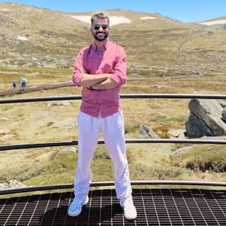 A man in a pink shirt and white pants stands with his arms folded on an iron platform with a dry, hilly terrain in the background.