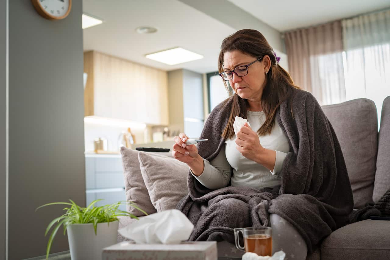 A woman sitting on sofa at home checking her temperature and holding a tissue.