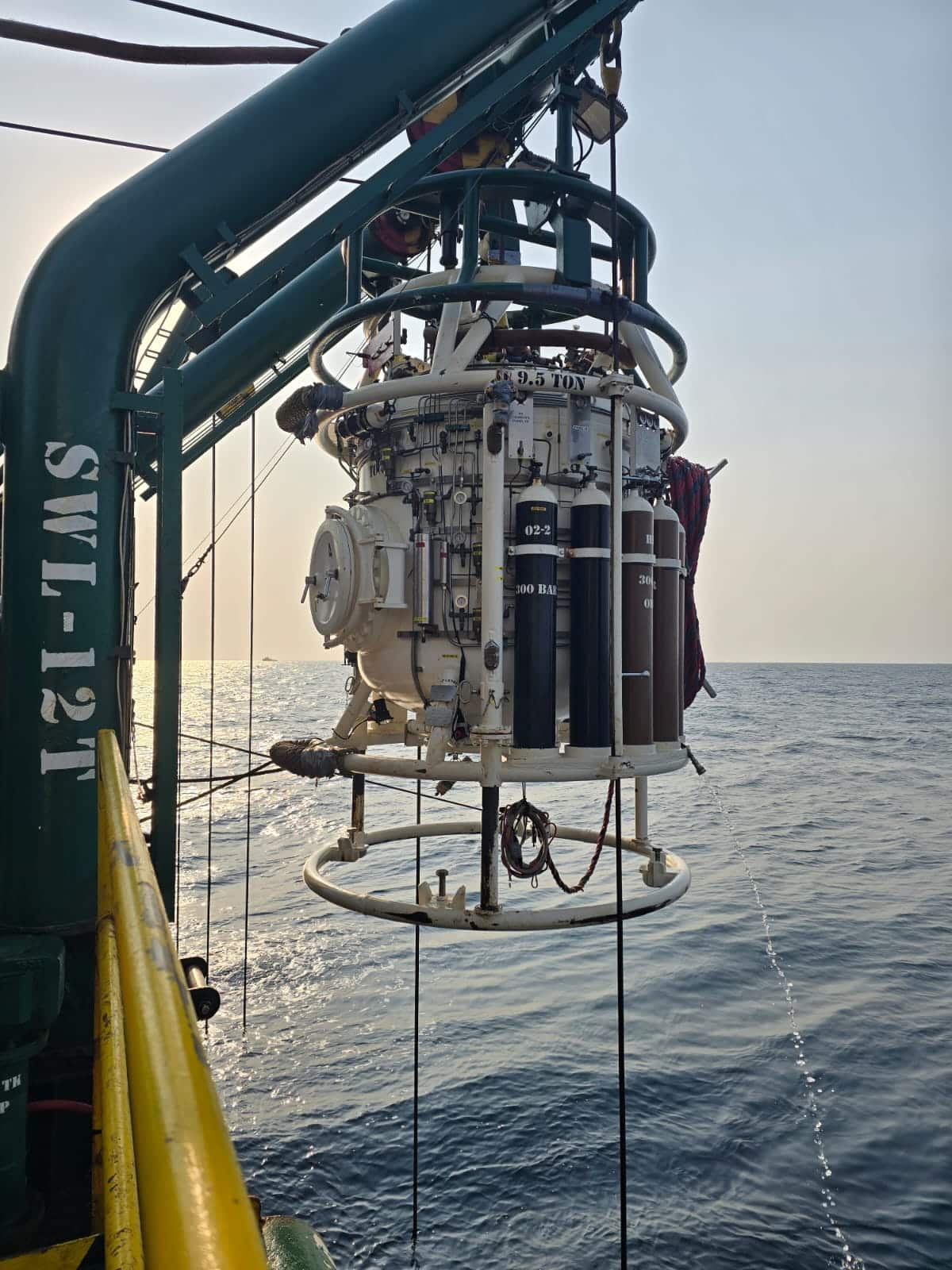 A diving bell is suspended in the air, ready to descend into the ocean carrying commercial divers.