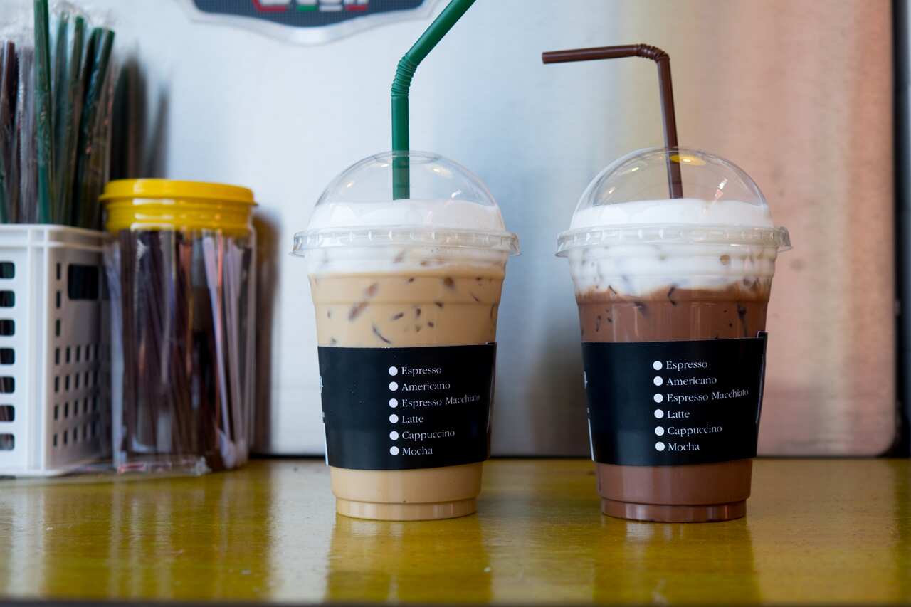 Two iced drinks in plastic drinking containers on a cafe bench.