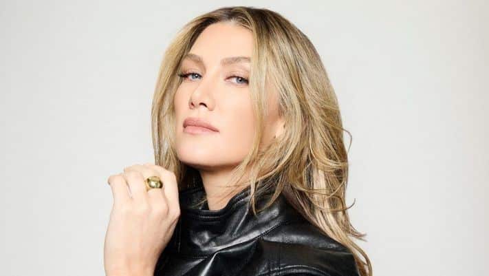 A blonde woman with blue eyes wearing a black leather jacket and a gold ring poses against a plain white background.