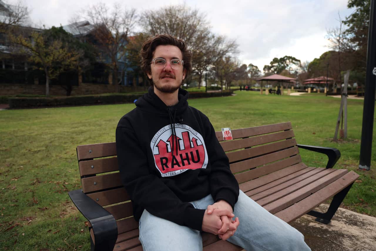 A young man wearing a black hoodie with glasses sat on a bench in the park, posing for a camera.