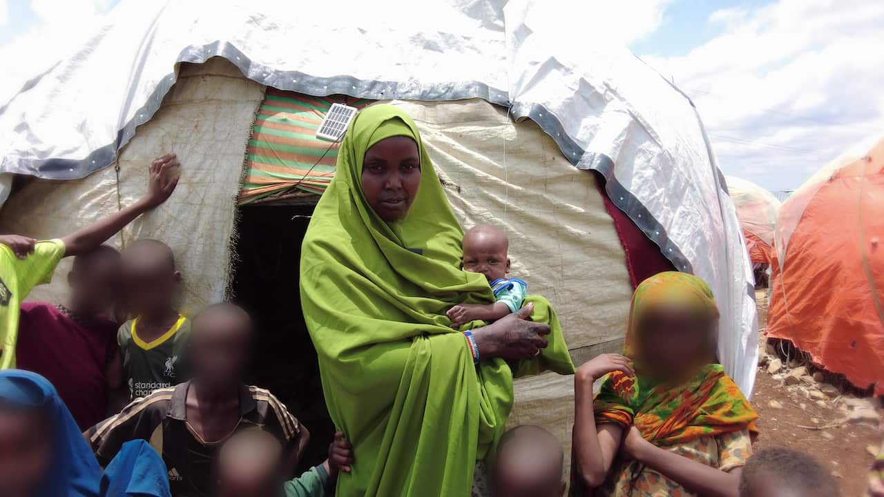 A woman standing outside a tent holding her child. Other children are standing around them.