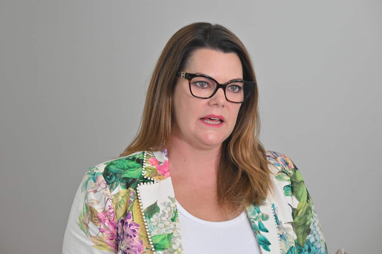 A woman in a floral blazer and glasses speaking in a press conference