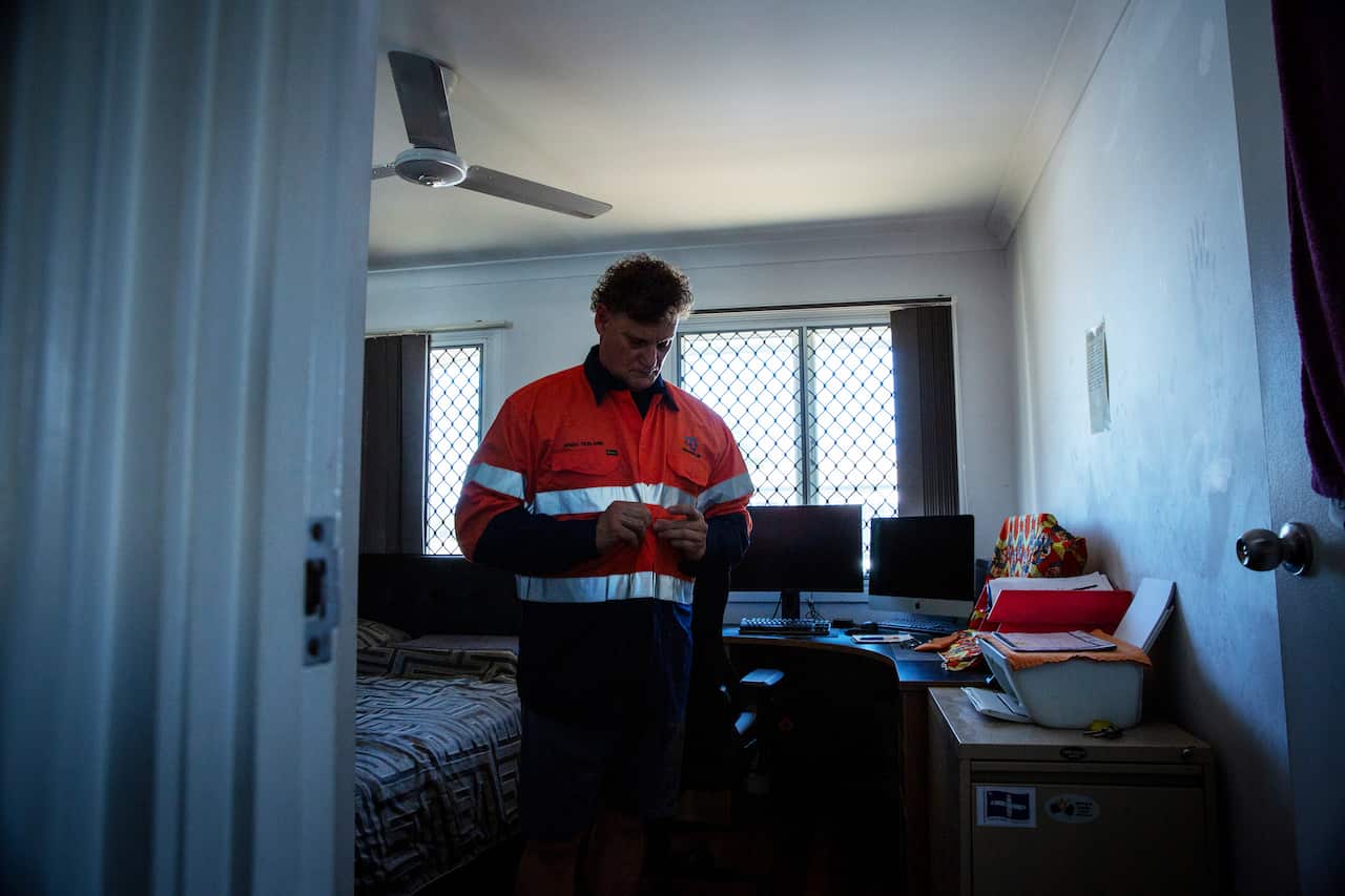 A middle-aged man wearing an orange hi-vis construction shirt stands in an unadorned bedroom.