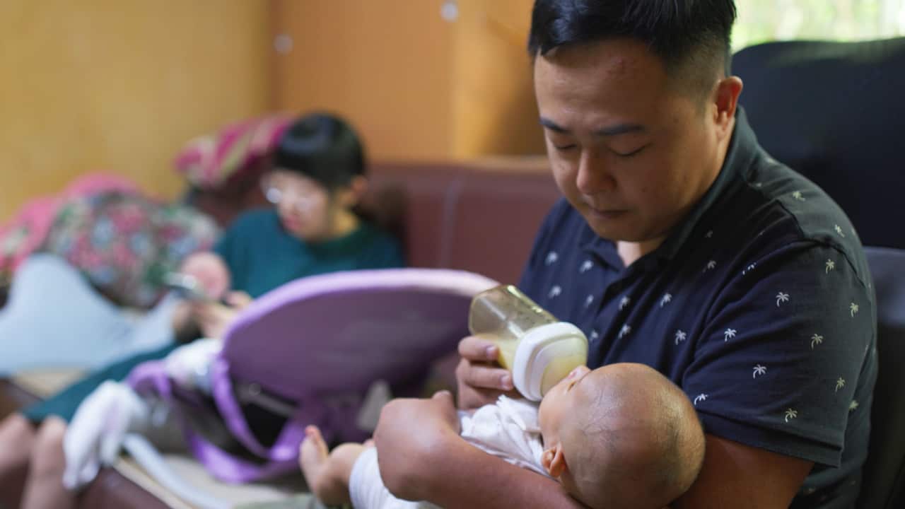 A man in a dark-blue polo shirt is holding and bottle-feeding a baby while a woman in the background is looking at her phone