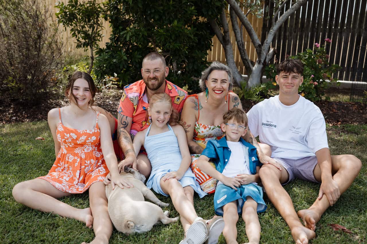 a family of two adults - a man and woman - with four young kids and a dog sitting on lawn smiling