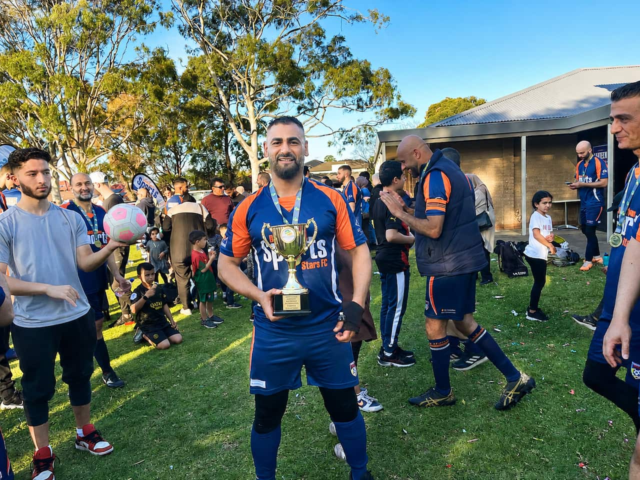 A man wearing a blue and orange football jersey smiles and holds up a golden trophy. Others in the same colours are around him.
