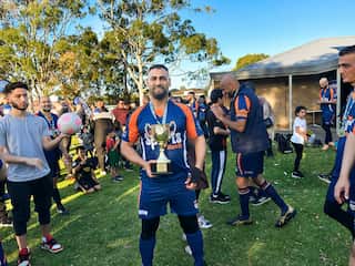 A man wearing a blue and orange football jersey smiles and holds up a golden trophy. Others in the same colours are around him.