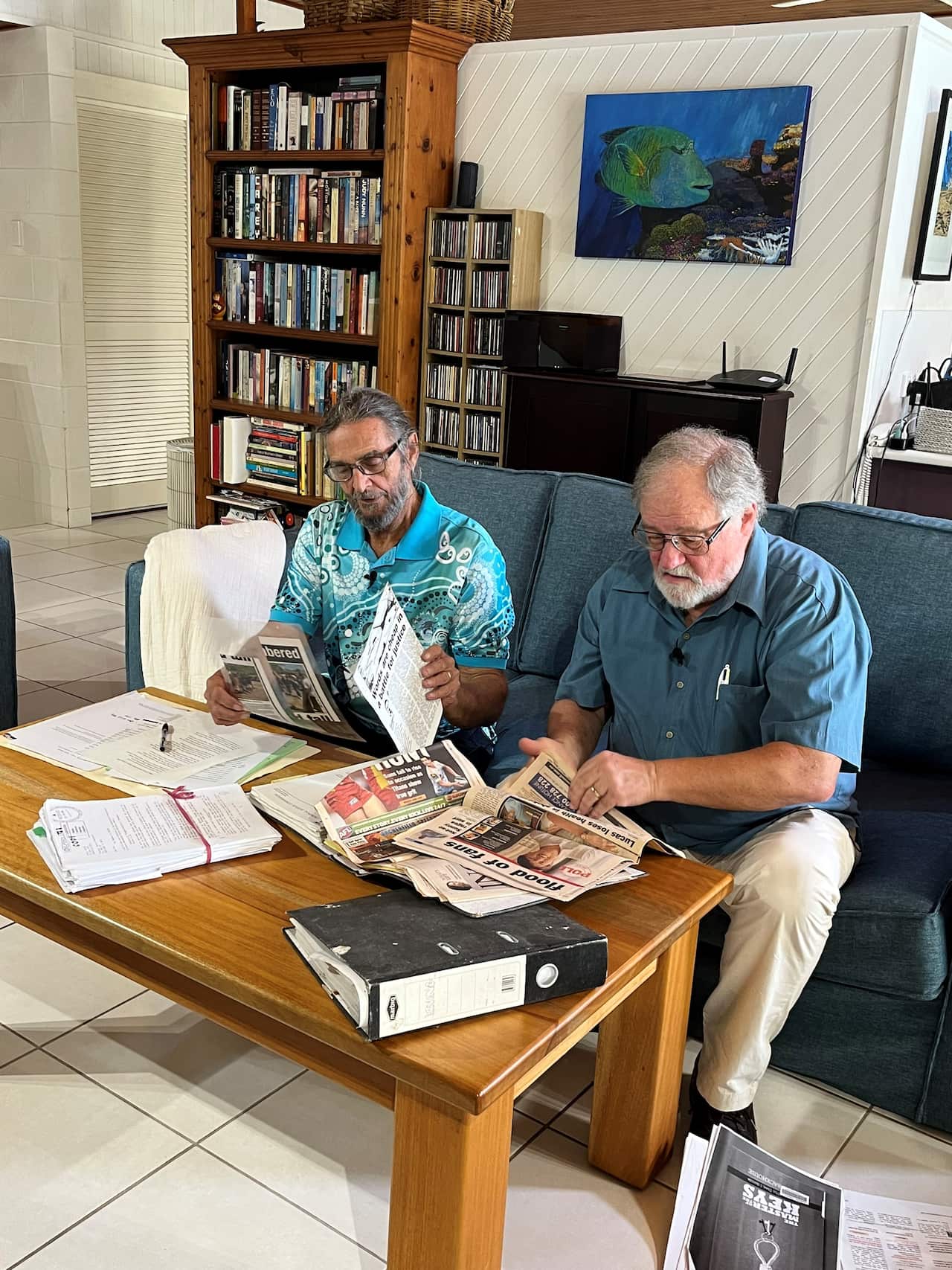 Two men with grey hair and glasses sit on a sofa in front of a coffee table where they are reading through papers and news reports.