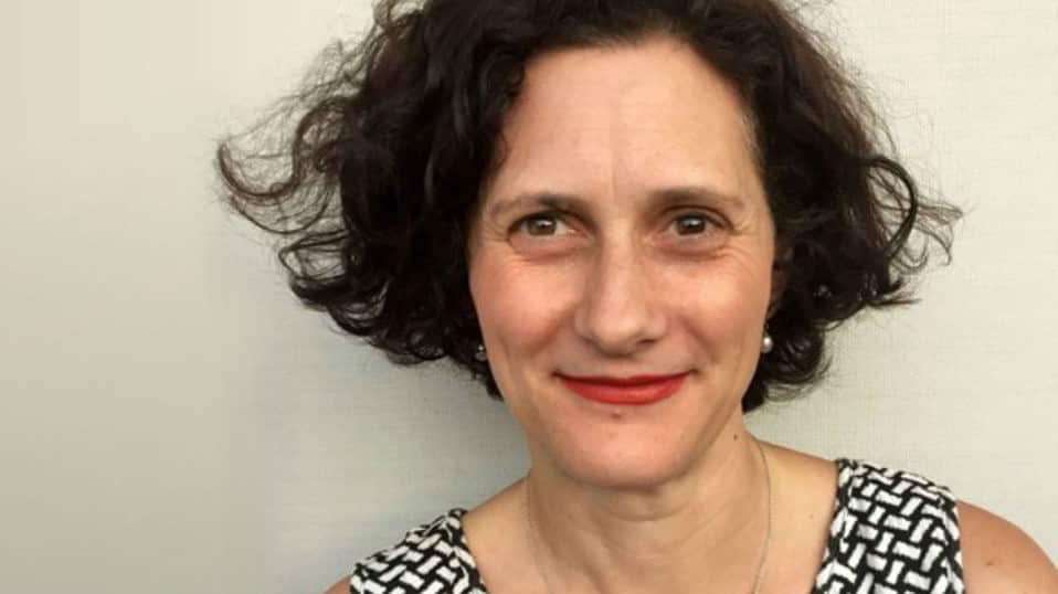 A woman with short, dark, curly hair is smiling.